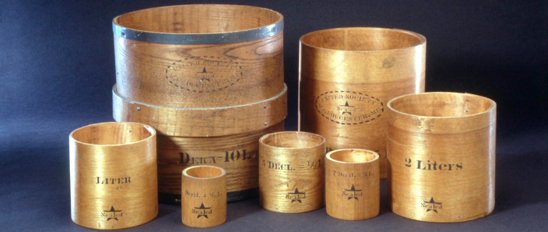 A set of seven copper-soldered wooden volumetric measures from largest to smallest, 1 dekaliter (a dekaliter is 10 liters), 1/2 dekaliter (5 liters), 2 liters, 1 liter, 5 deciliters (a deciliter is 1/10 of a liter or 100 cubic centimeters), 2 deciliters, and 1 deciliter. 